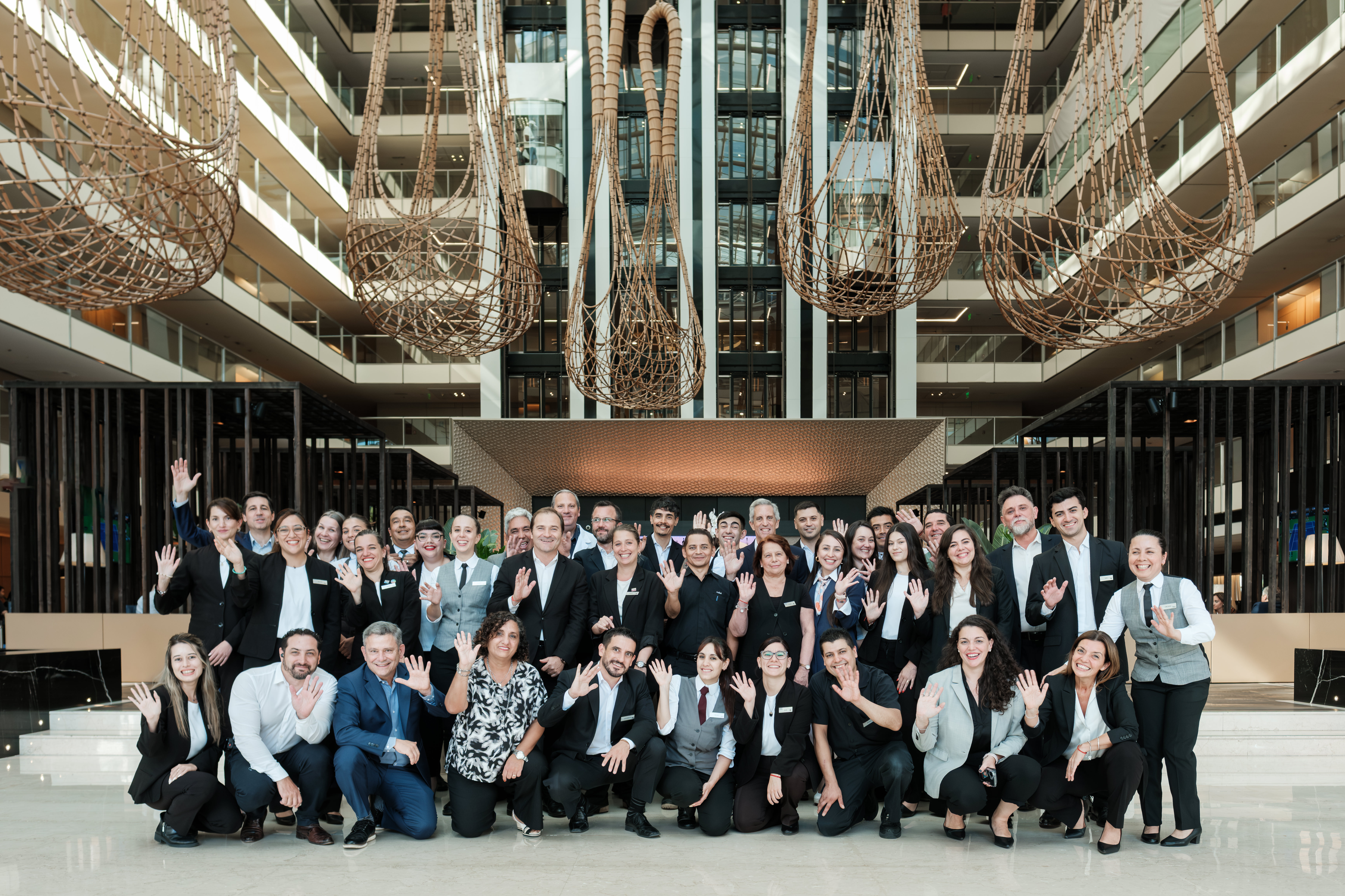 Large group Hilton Team Members in Argentina posing and waving inside hotel lobby with large hanging wicker light fixtures.