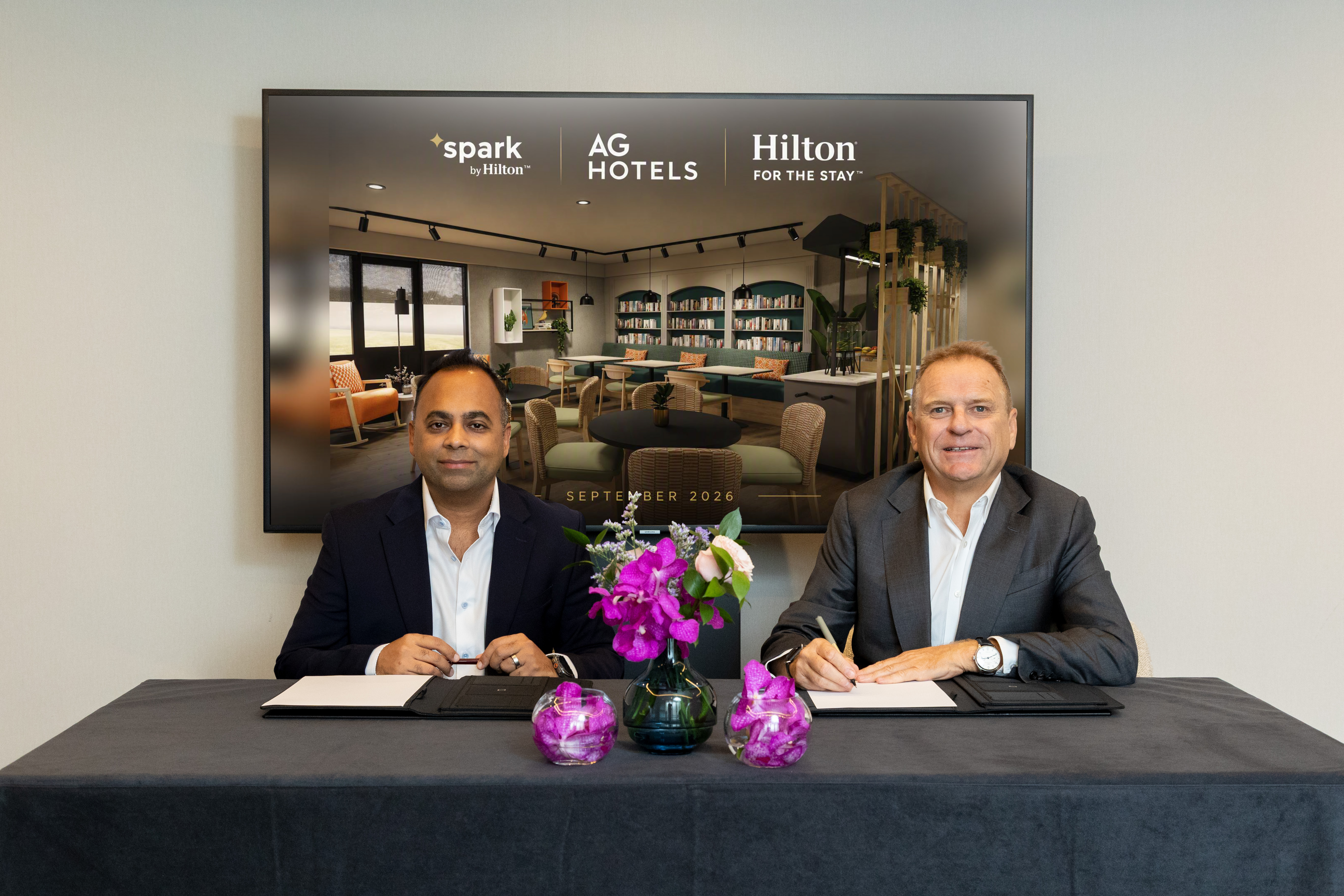 Two men in suits sit at a table signing AG Hotels Group and Hilton Signing Announcement, with a floral centerpiece and a screen behind them displaying Hilton and AG Hotels logos.