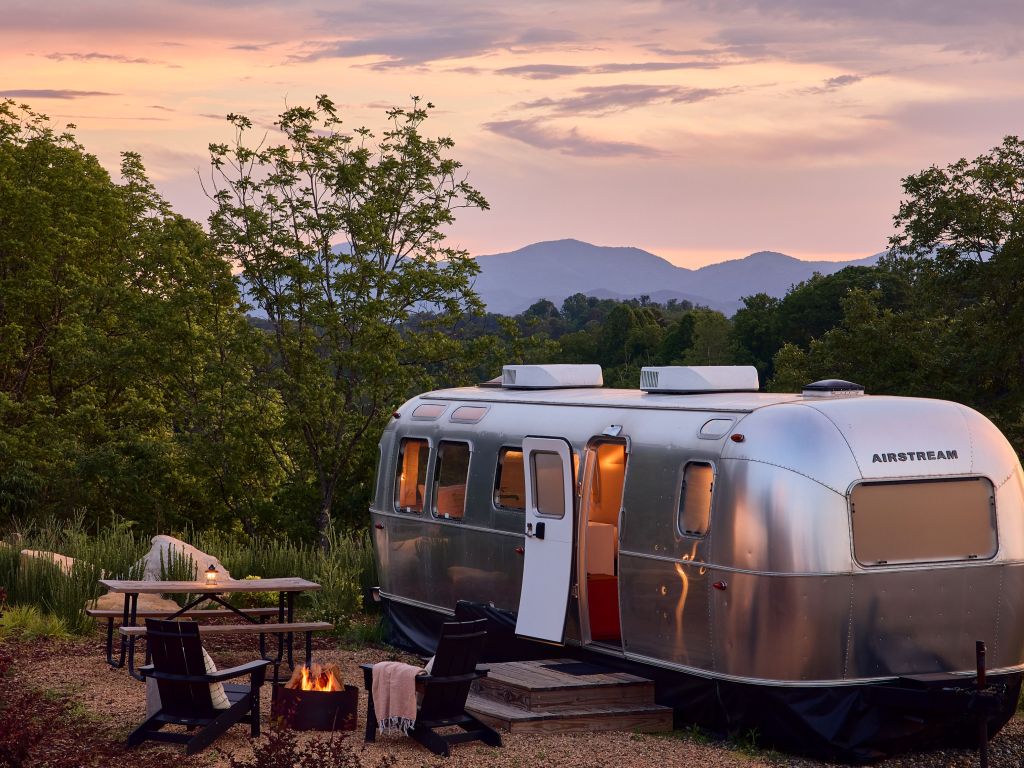 A silver Airstream at AutoCamp Asheville with an open door is set in a wooded area at sunset, with two chairs around a fire pit and a picnic table nearby.
