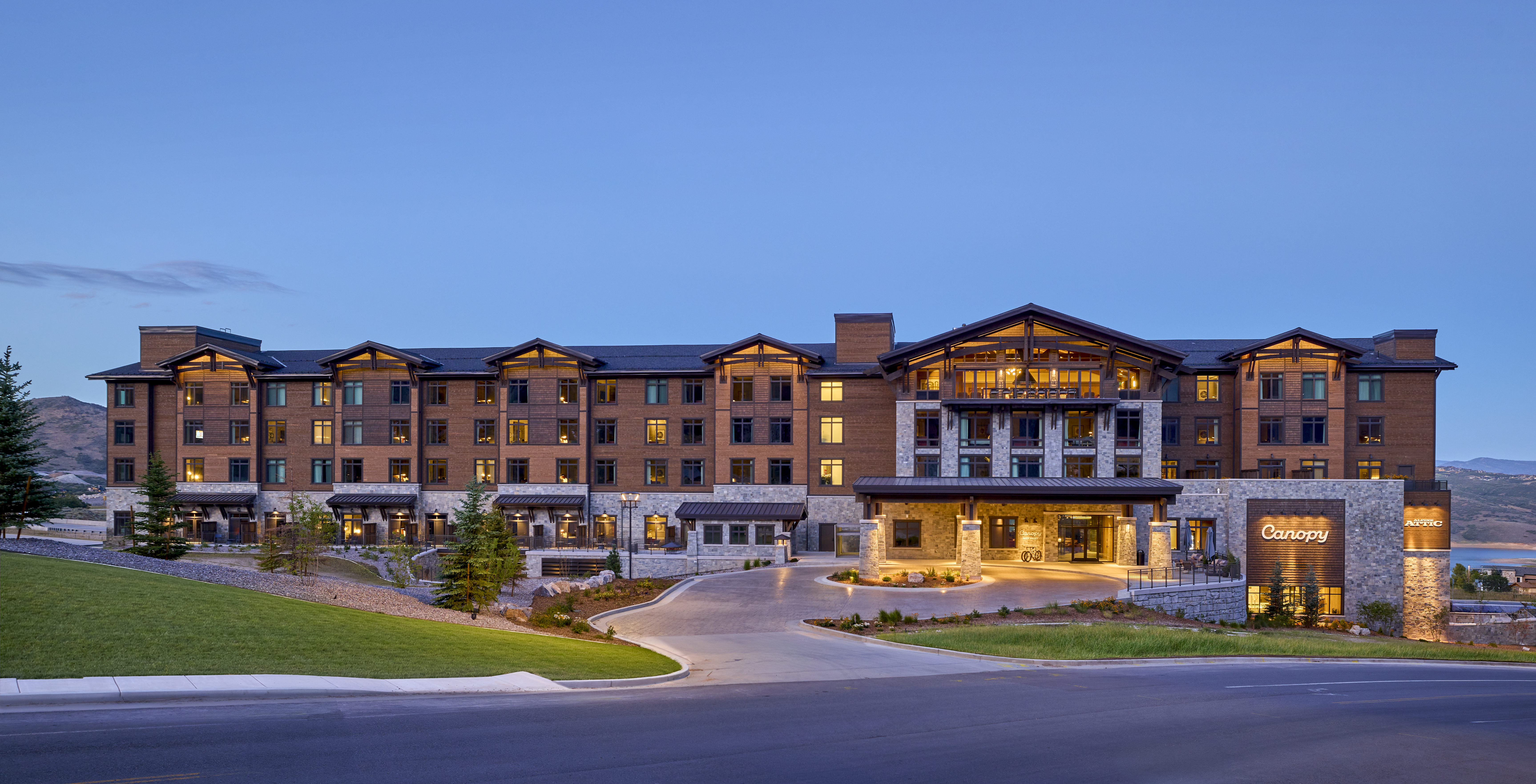 Large modern hotel building at Canopy Deer Valley with warm interior lights and a driveway entrance, surrounded by landscaped greenery.
