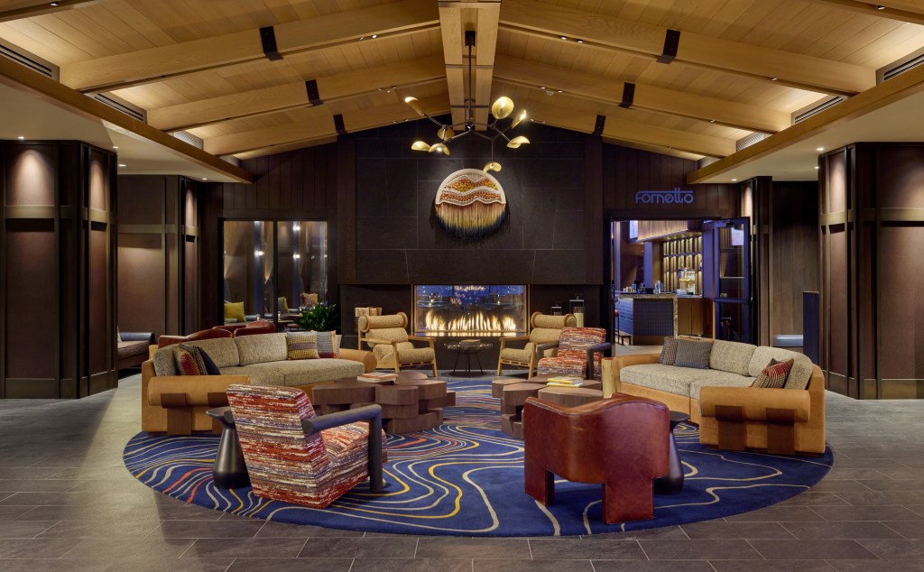 Luxurious lounge area with modern sofas and armchairs arranged around a fireplace, featuring a blue patterned rug and wooden ceiling at Canopy Deer Valley.