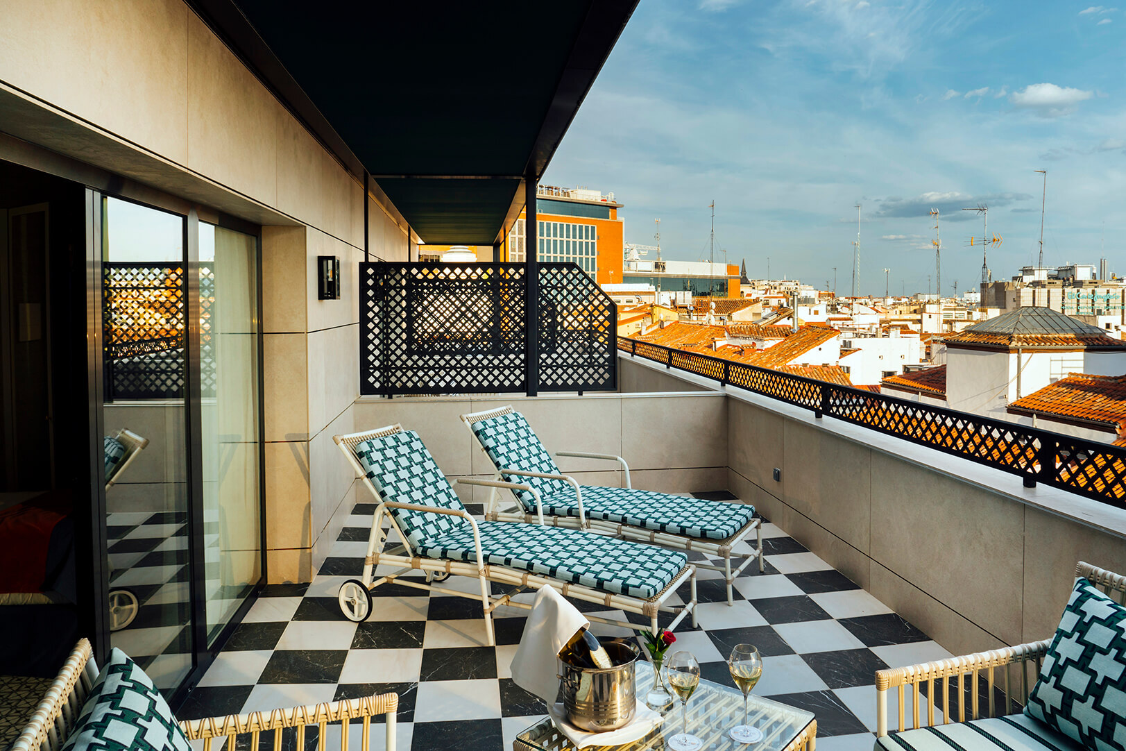 Casa Verbena Madrid, Outset Collection by Hilton balcony with checkered floor, two lounge chairs, a small table with wine glasses and a cityscape view under a blue sky.