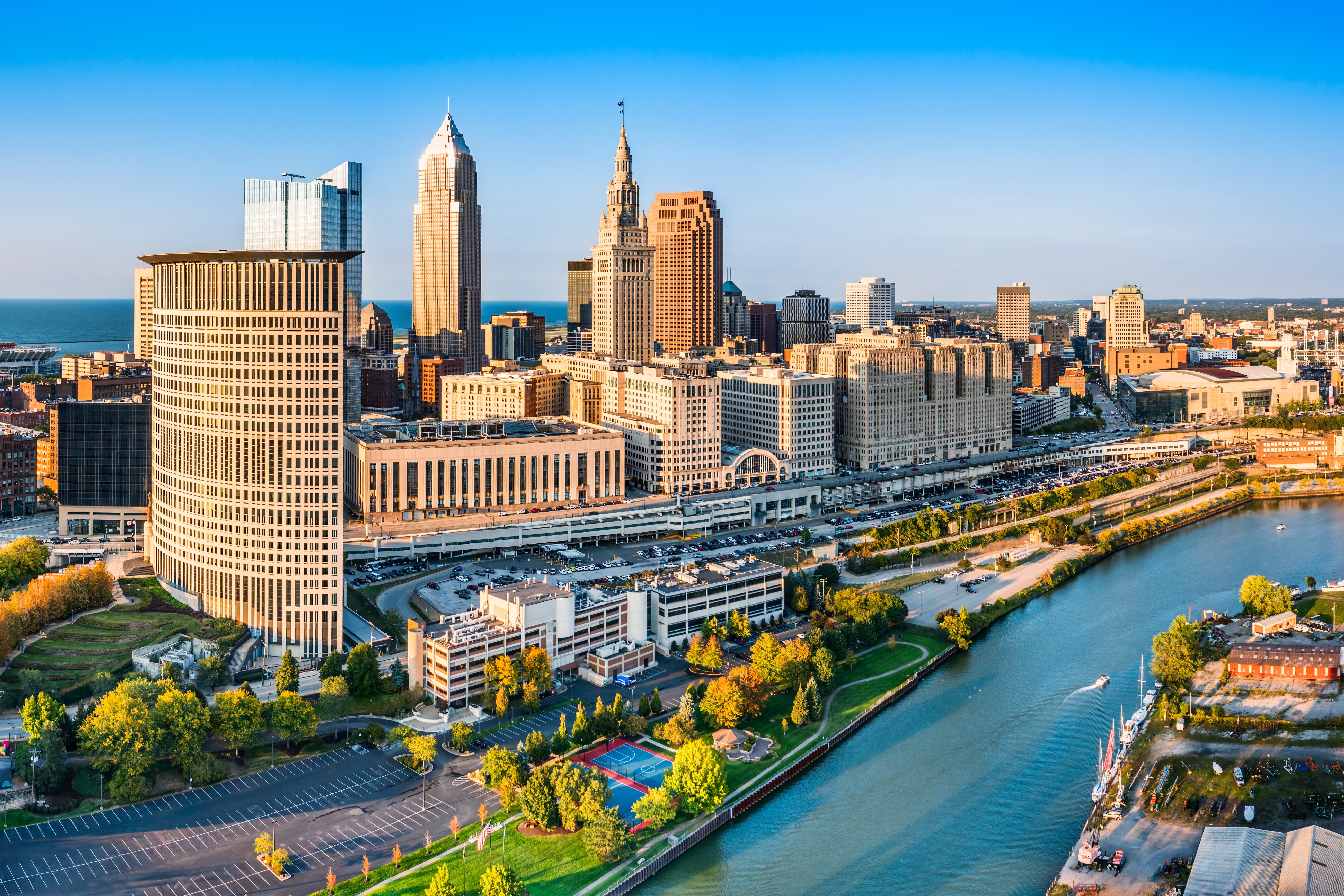 Downtown Cleveland skyline with tall buildings, a riverfront park, and boats on the water under a clear blue sky.