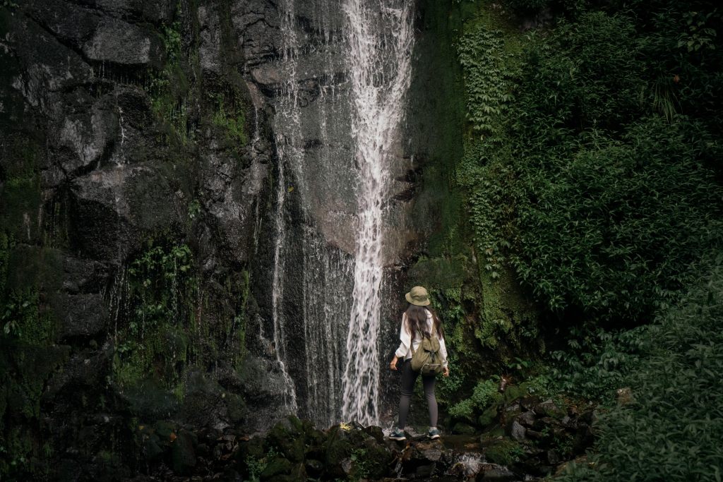 Person wearing a hat and backpack standing on rocks, facing a narrow waterfall surrounded by moss-covered rocks and dense greenery at Conrad Bali.