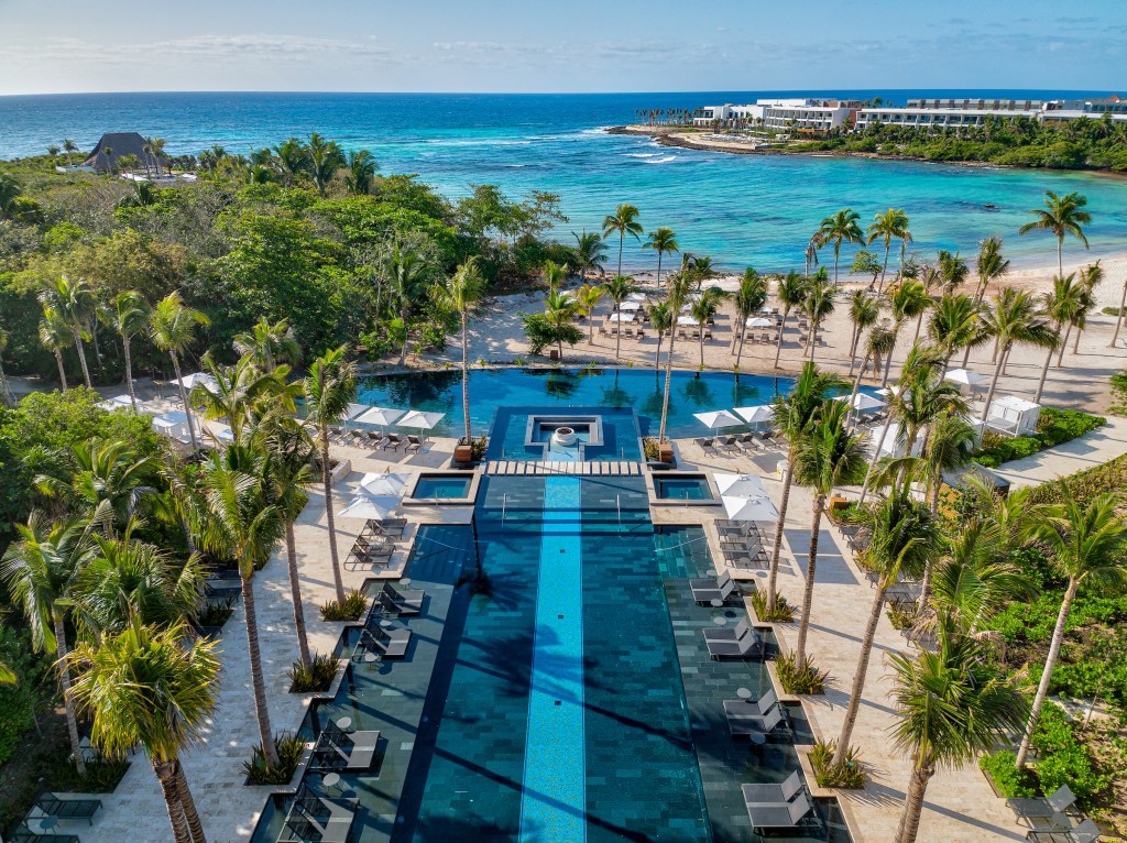 Conrad Tulum Riviera Maya luxury resort pool area with palm trees, lounge chairs, umbrellas, and a view of the ocean and sandy beach.