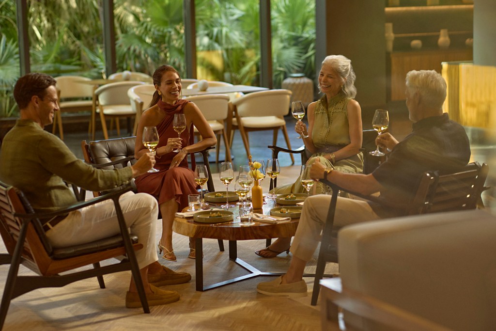 Four adults sitting around a low wooden table, enjoying drinks and conversation in a cozy, plant-filled indoor setting at Conrad Tulum Riviera Maya.