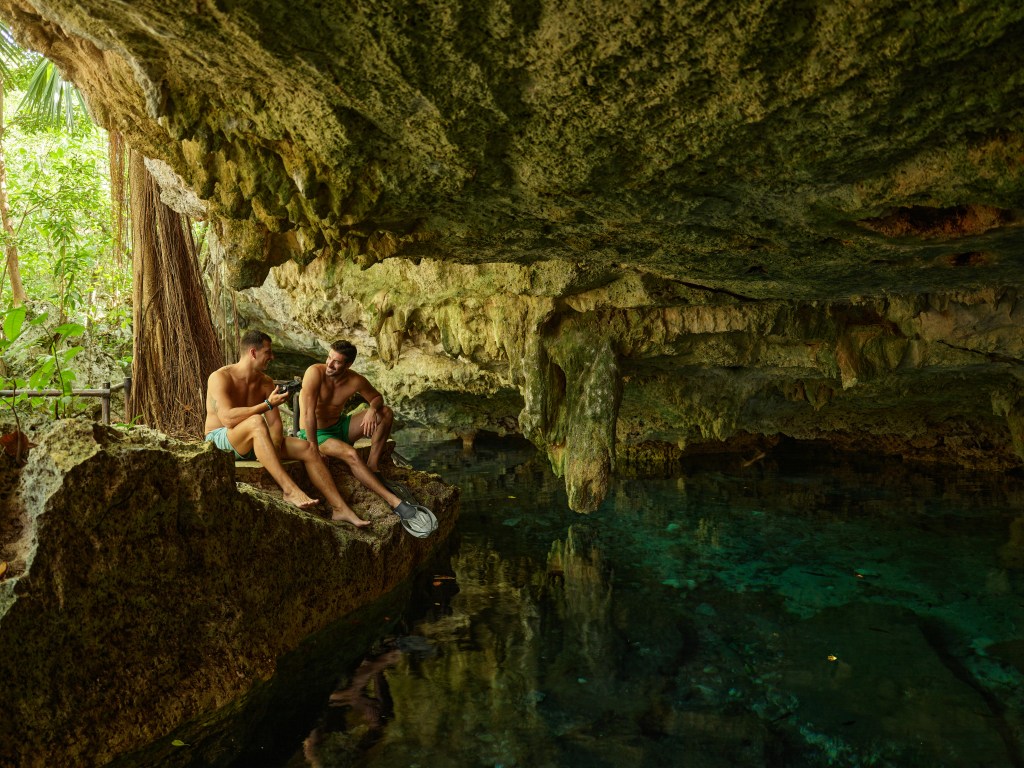 Two men in swim trunks sitting on a rock ledge inside a cave with clear water below, surrounded by natural rock formations and greenery at Conrad Tulum Riviera Maya Cenote Excursion.