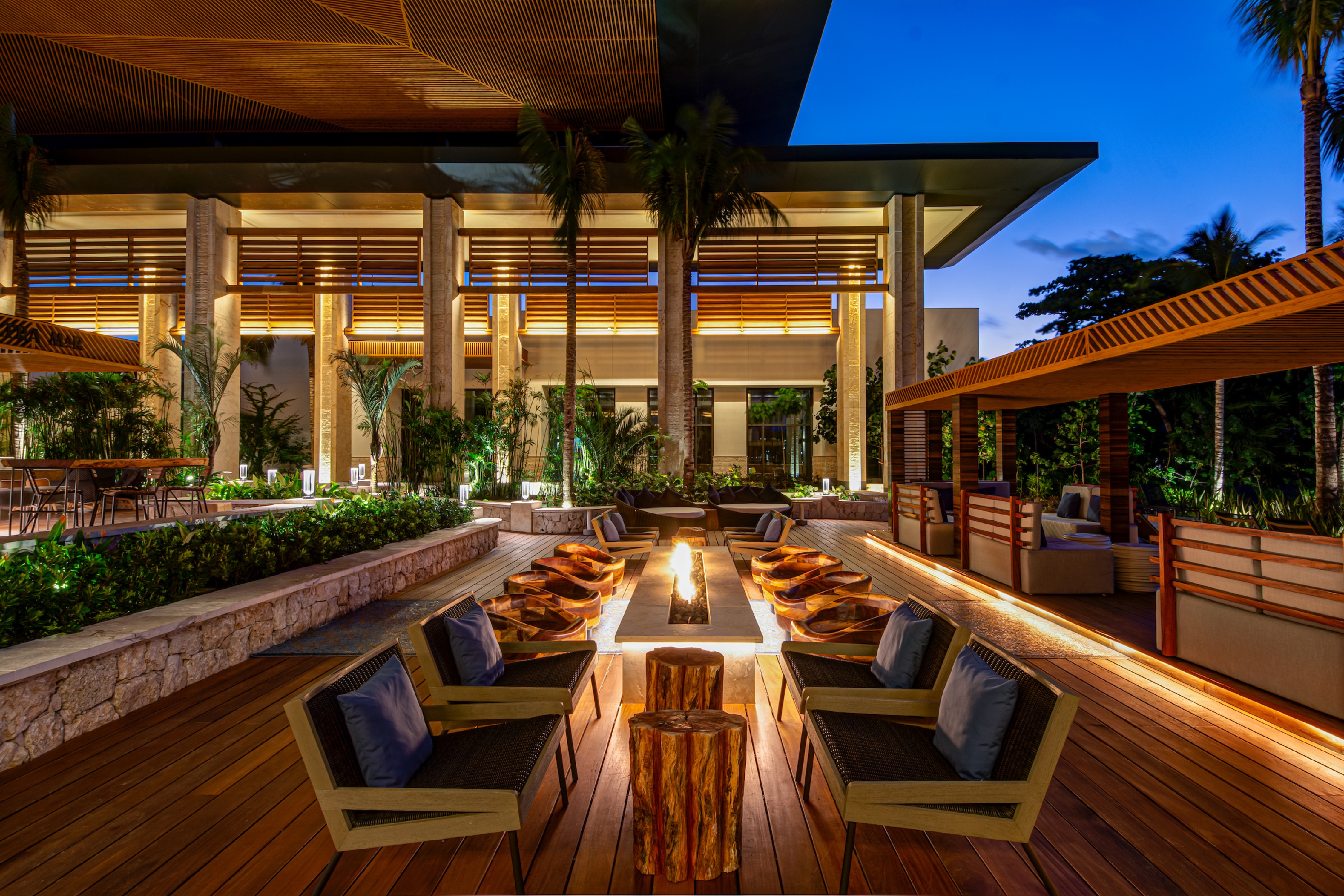 Outdoor seating area with modern chairs and a central fire pit, surrounded by wooden decking and illuminated architectural elements at Conrad Tulum Riviera Maya.