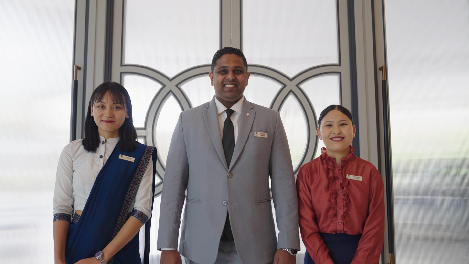 Three smiling professionals standing side by side in front of window, dressed in formal and traditional attire.