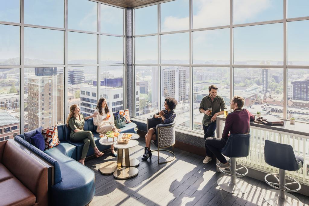 Five people socializing in a bright lounge with large windows overlooking a cityscape, enjoying drinks and snacks at Embassy Suites by Hilton.