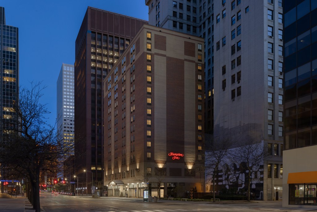 Downtown city street at dusk with Hampton Inn Cleveland-Downtown building lit with surrounding skyscrapers.