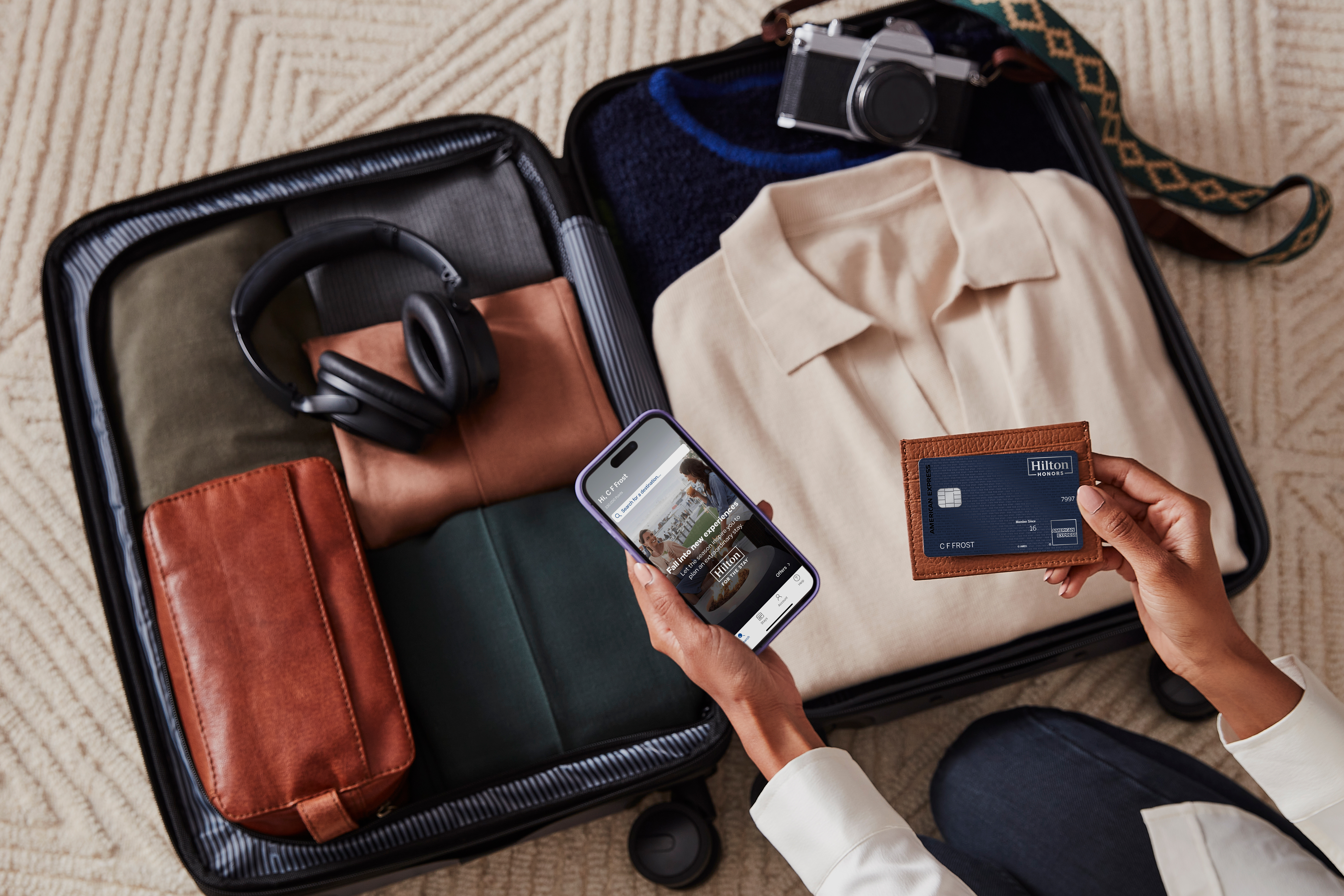 Person holding a Hilton Honors credit card and a phone over an open suitcase packed with clothes, headphones, and a camera.