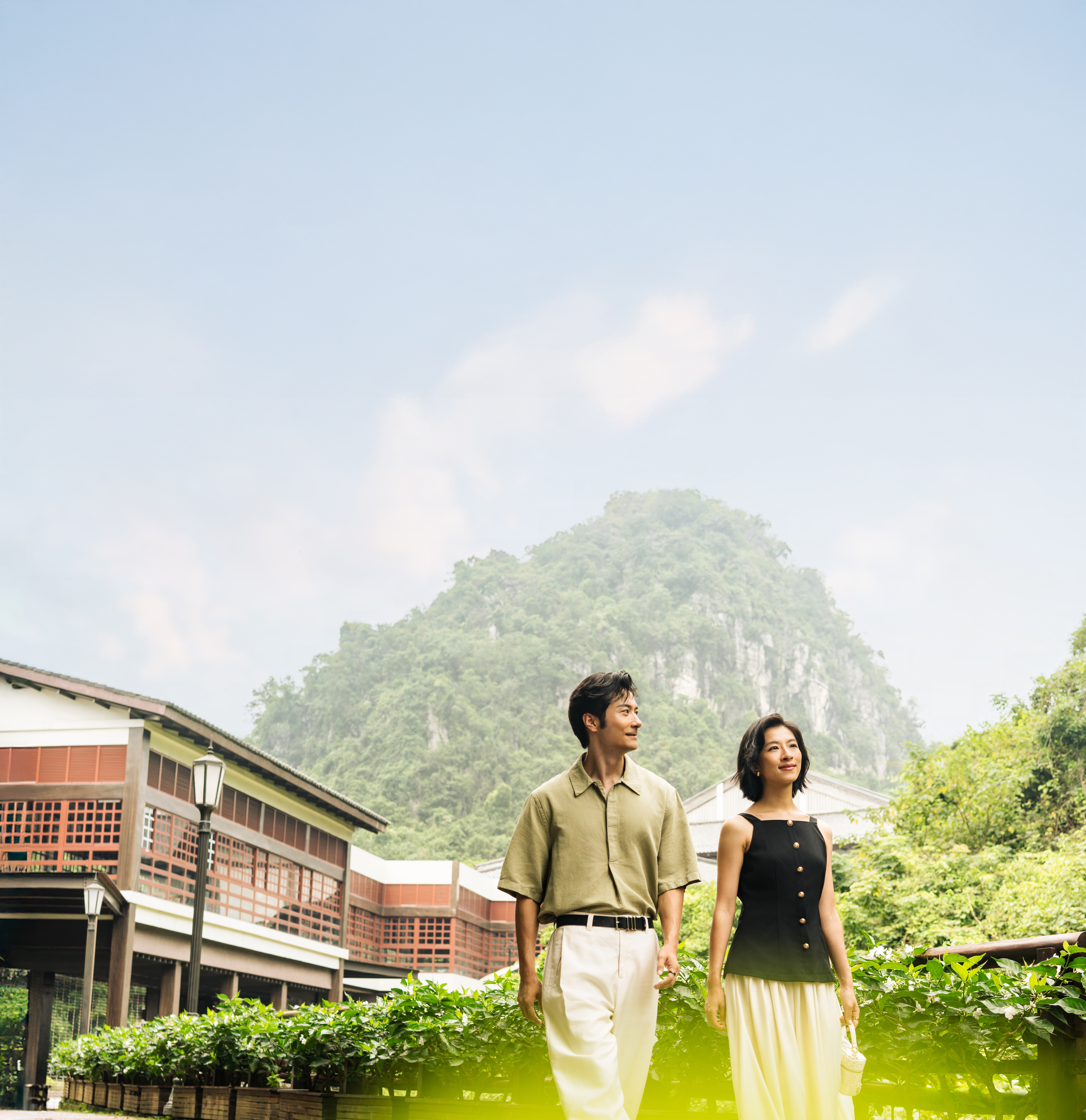 Couple walking outdoors near greenery and traditional buildings with a mountain in the background at Hilton Quang Hanh Onsen-Resort Grounds.