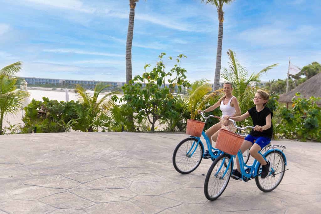 A woman and a boy riding blue bicycles with orange baskets on a paved area near palm trees and greenery under a blue sky.