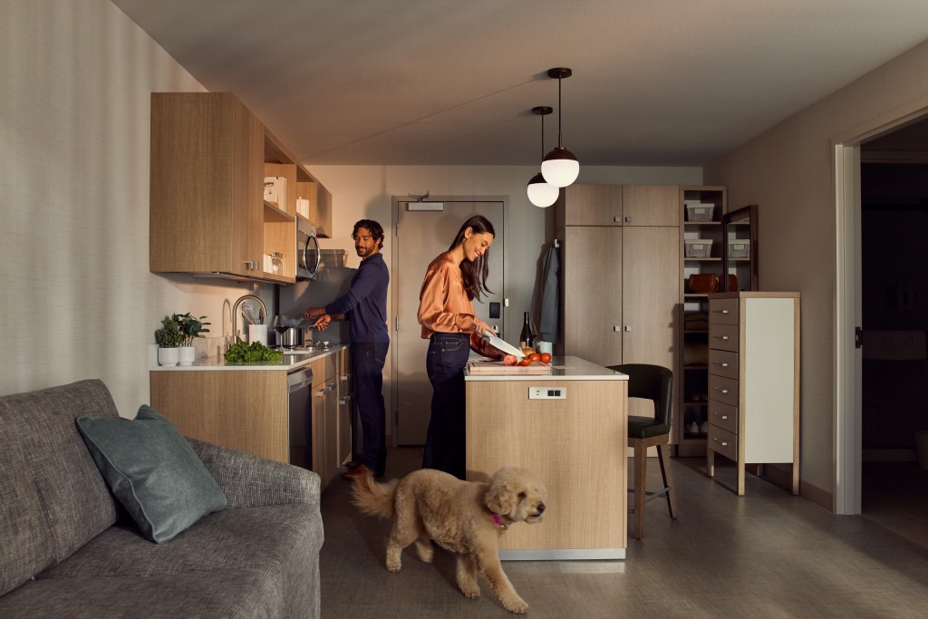 Couple cooking together in a Homewood Suites by Hilton In-Room modern kitchen with a dog walking in the foreground.