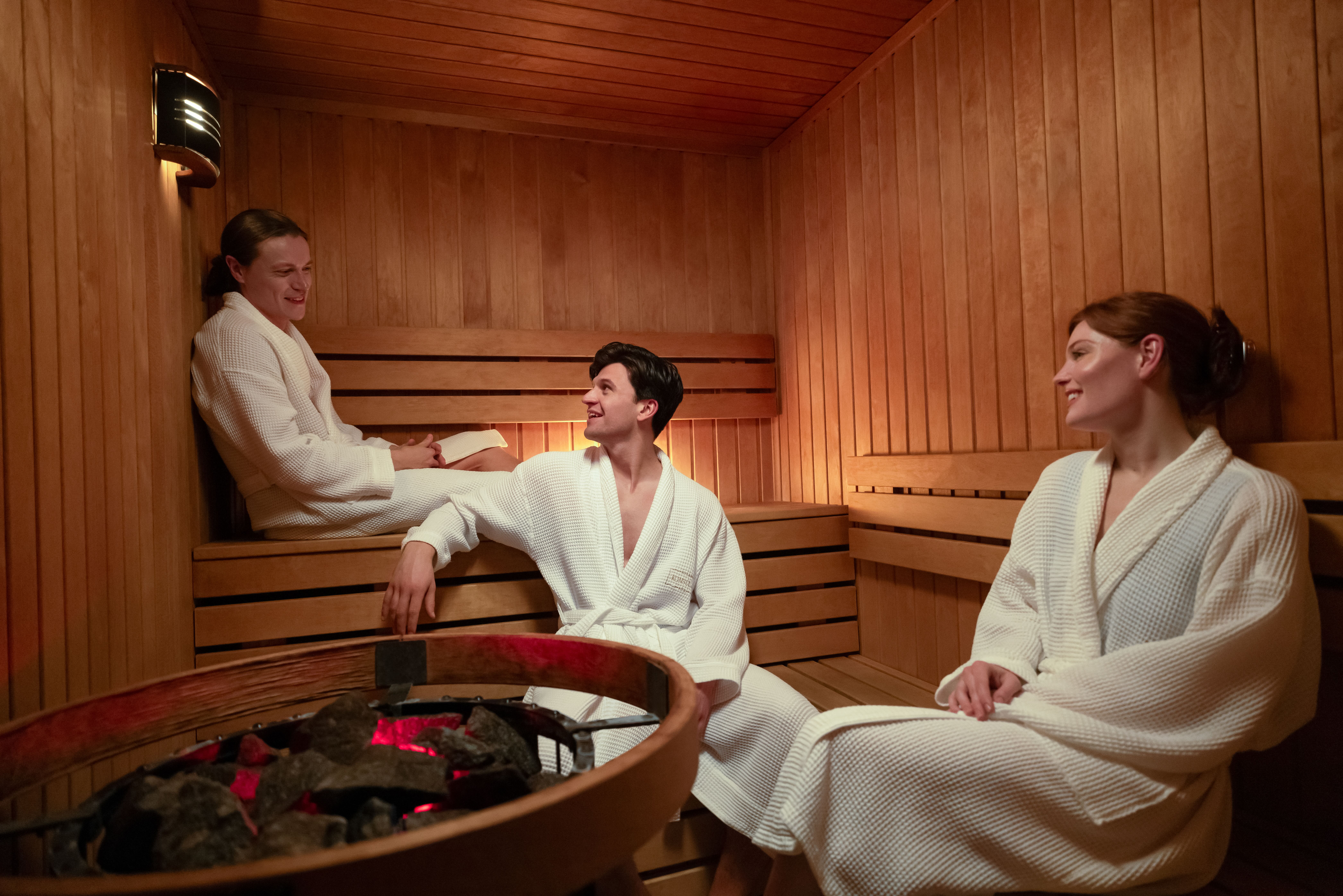 Three people in white robes relaxing and chatting inside a wooden sauna at Reykjavik Consulate Hotel, Curio Collection by Hilton.