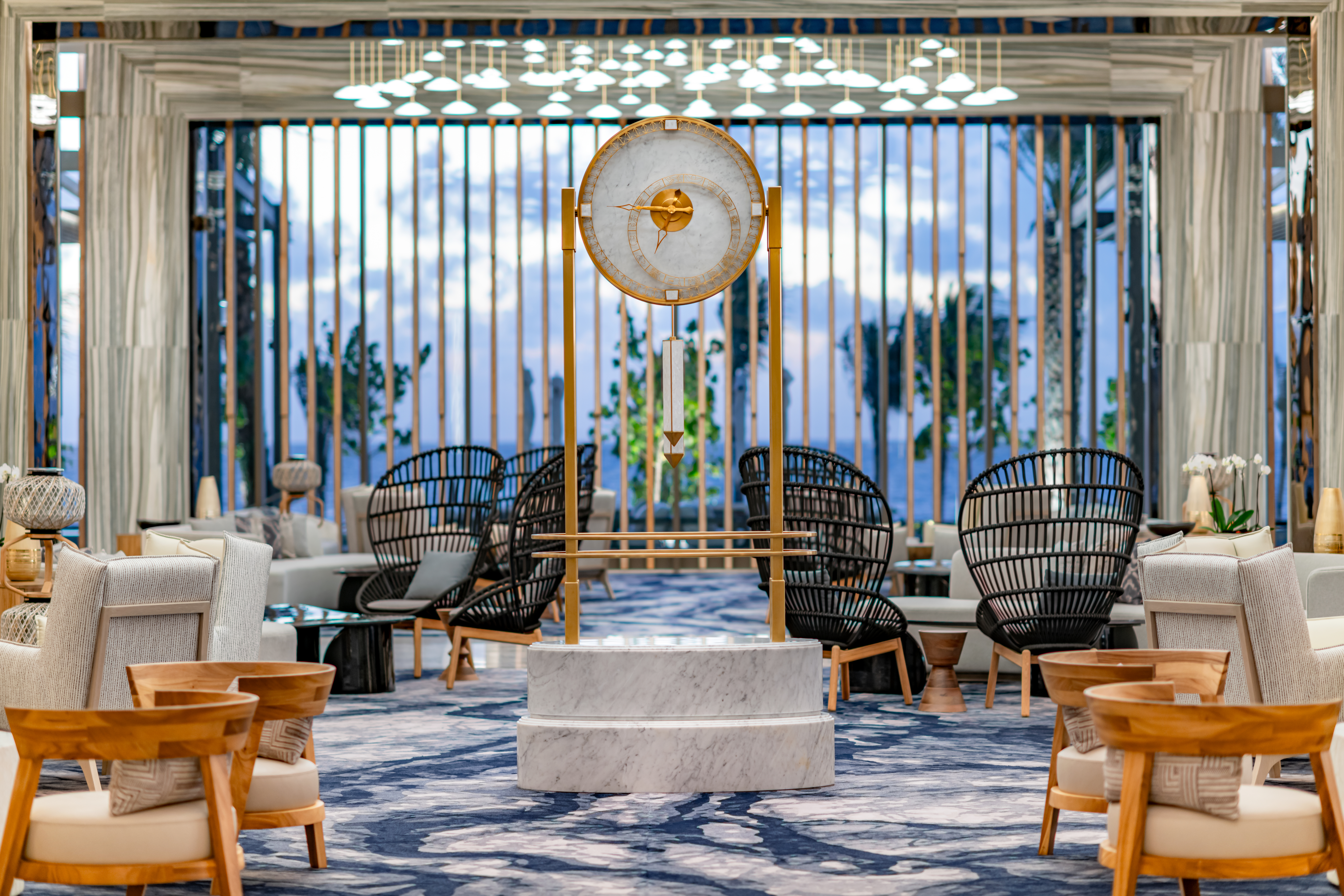 Modern lounge with stylish black and wooden chairs, a large decorative clock, and floor-to-ceiling windows with vertical bars at Waldorf Astoria Riviera Maya.