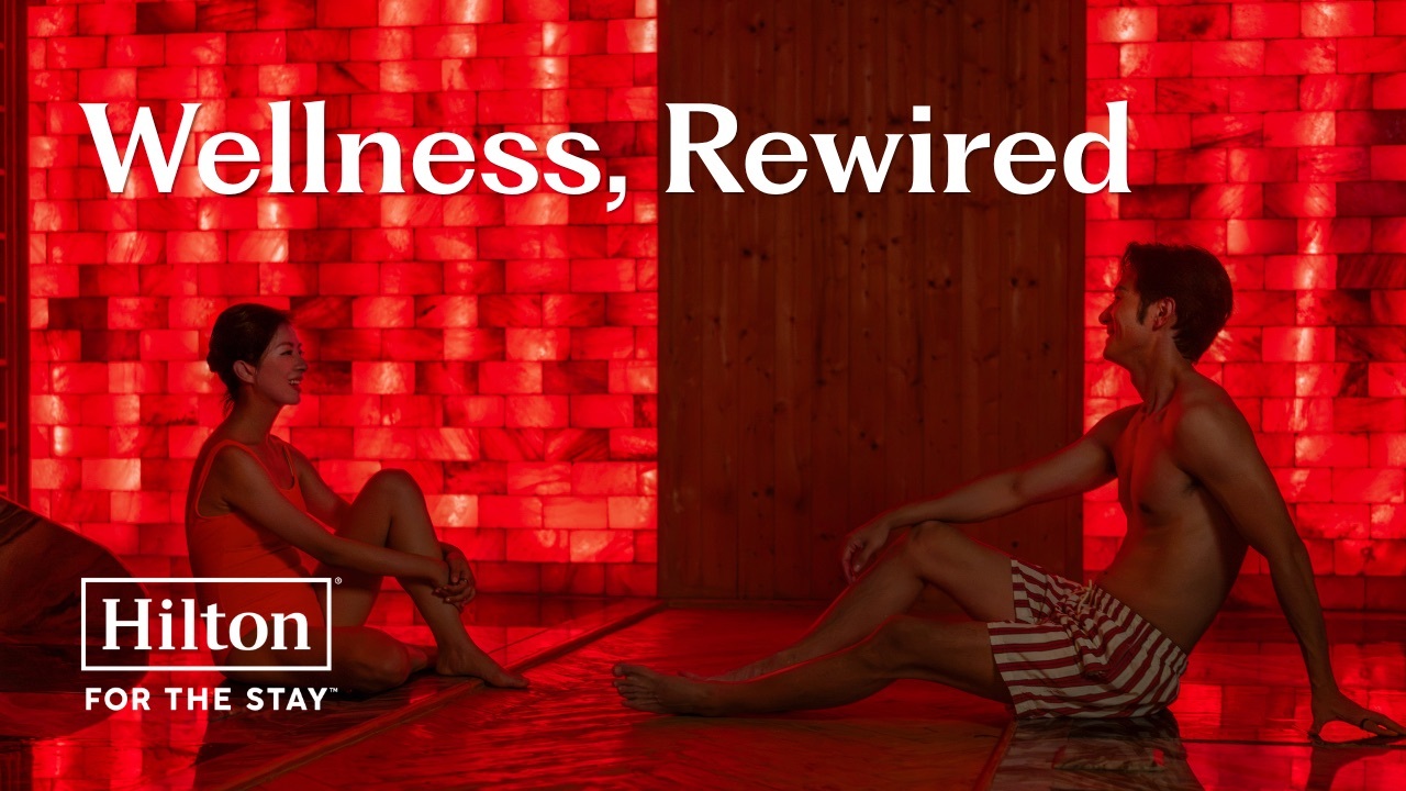 Two people sitting and relaxing in a red-lit sauna with the text "Wellness, Rewired" and the Hilton logo.