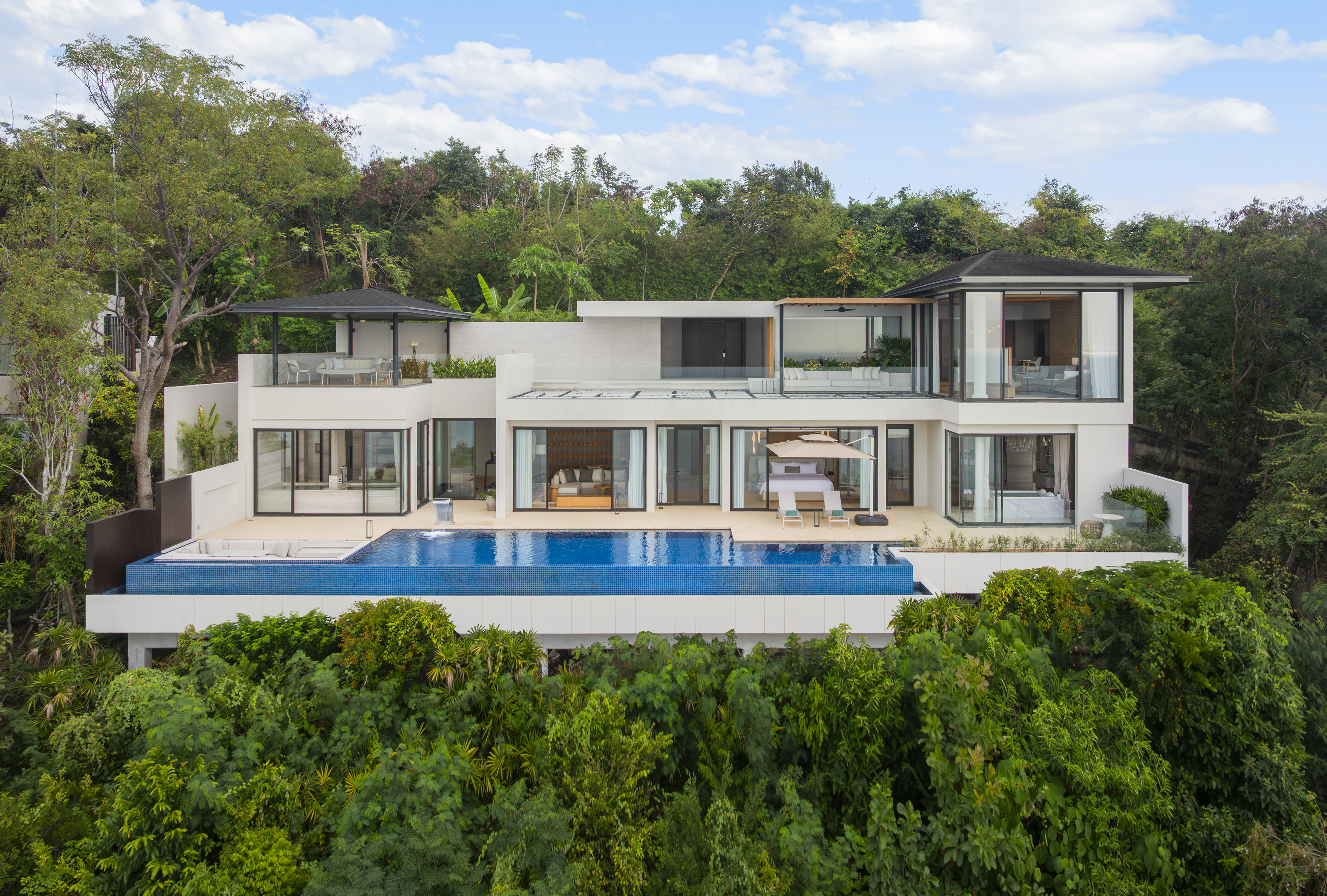 The Pearl Exterior Modern white villa at Conrad Koh Samui with large glass windows, an infinity pool, surrounded by lush green trees.