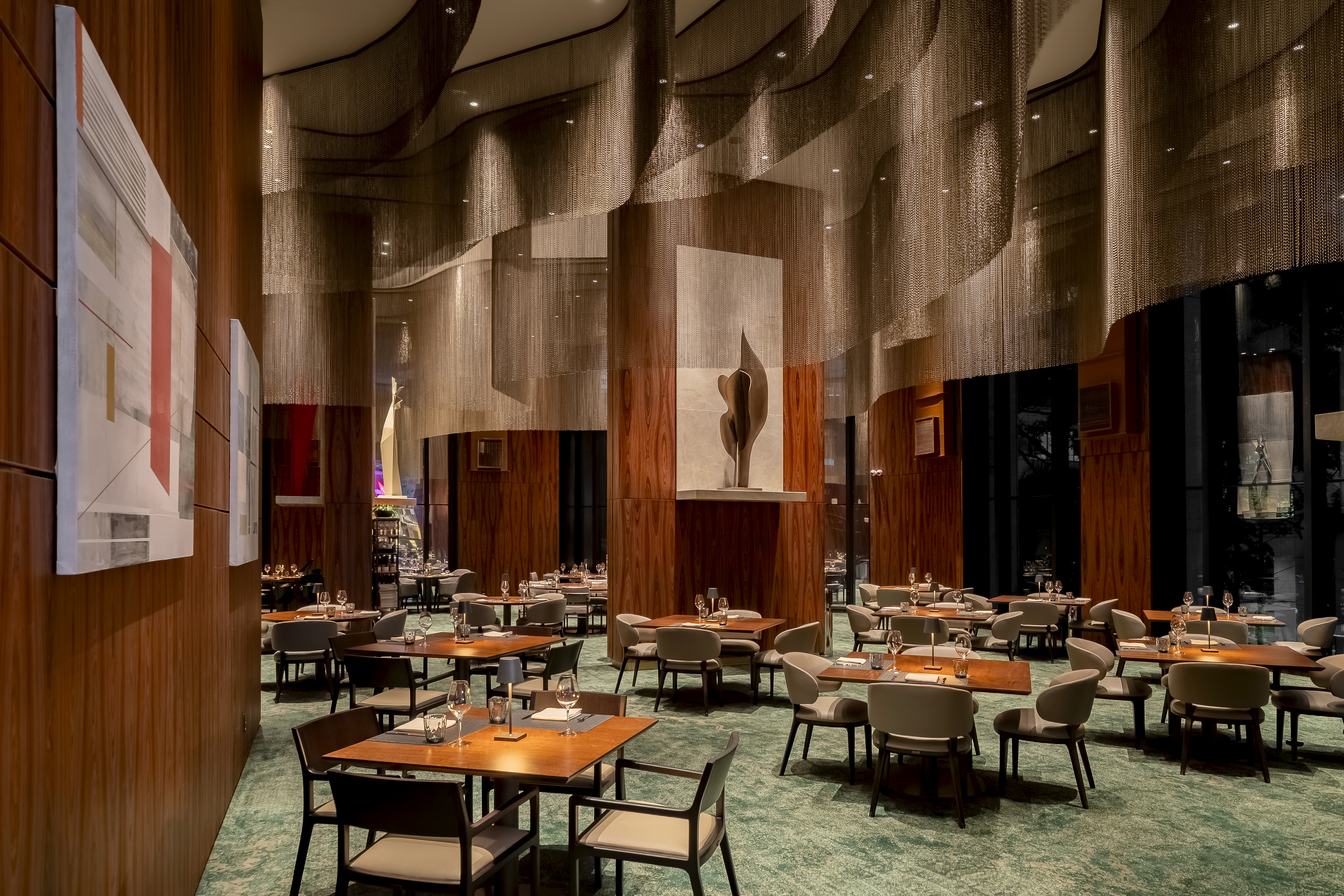Upscale restaurant interior at Hilton Sao Paulo Morumbi with wooden tables, beige chairs, green carpet, abstract wall art, and a decorative sculpture under a flowing metallic ceiling installation.
