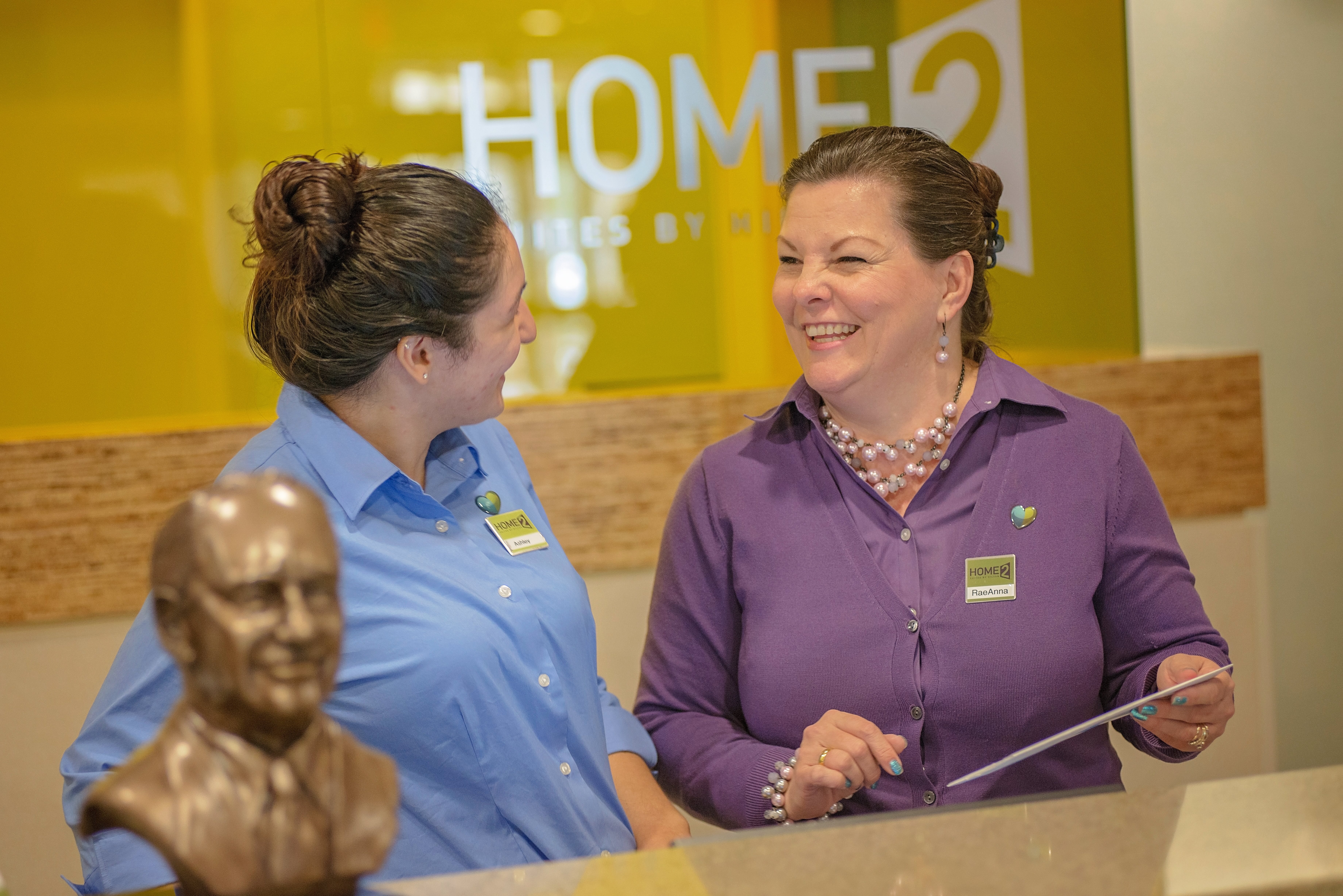 Two smiling women wearing name tags and uniforms are engaged in conversation behind a reception desk with a bronze bust in the foreground.