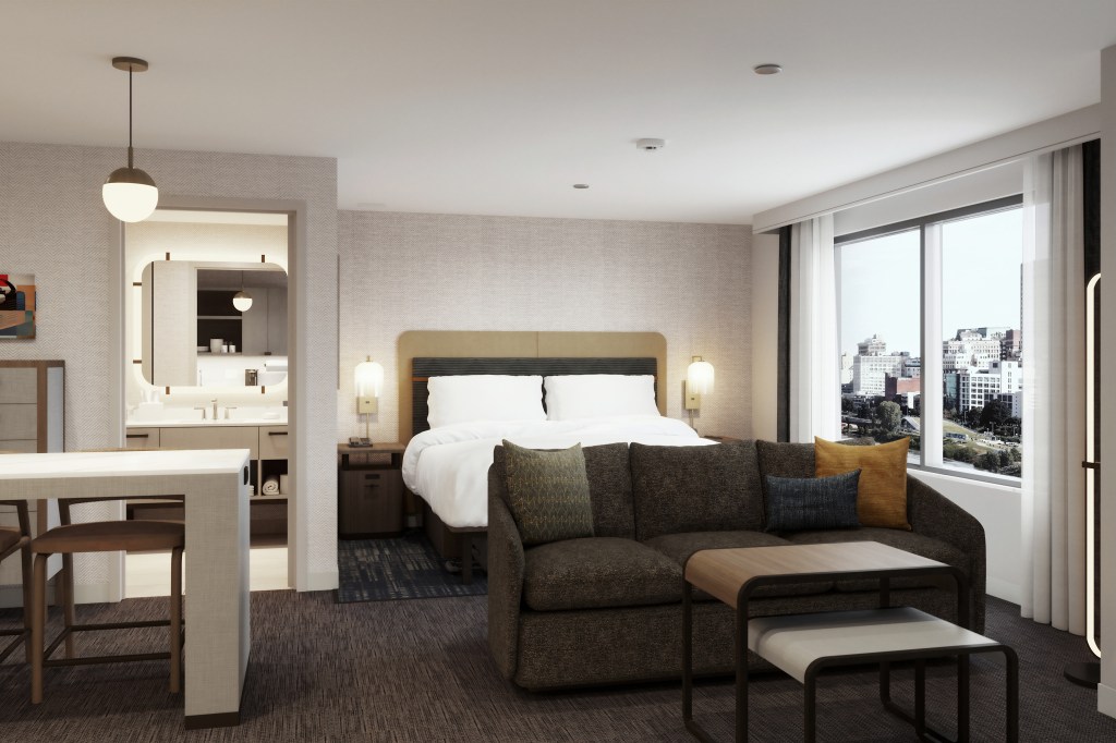 Modern hotel room with a large bed, dark sofa with colorful pillows, a small table, and a window showing a city view.