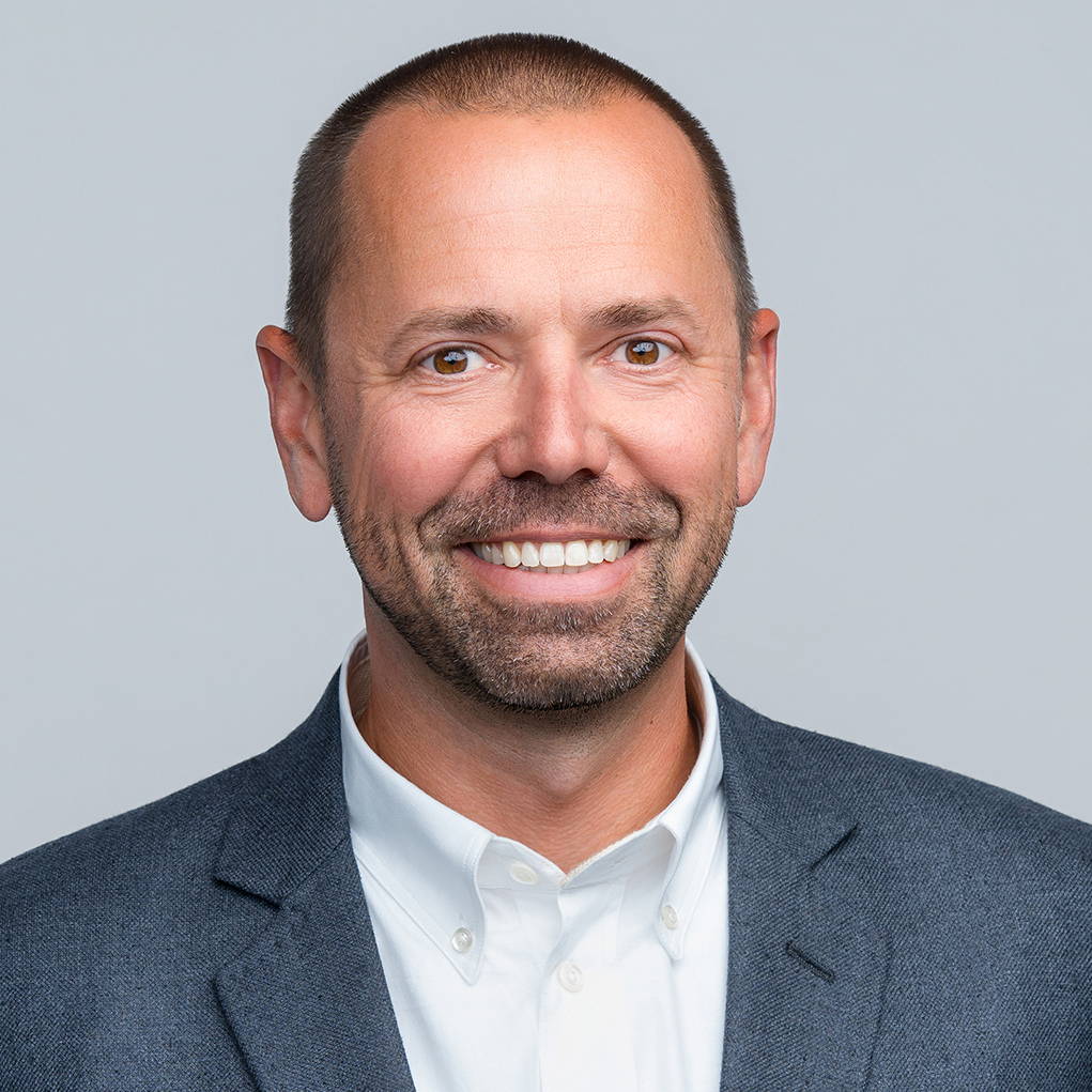 Jeff Moore Headshot -Smiling man with short hair and a beard, wearing a white shirt and a gray blazer against a light gray background.